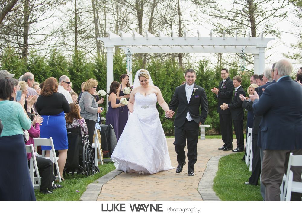 Newlyweds walk down a brick aisle, guests clap. White archway, green grass, trees.
