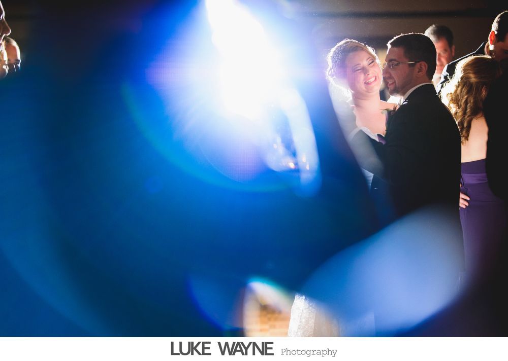 Bride and groom dancing at a wedding with a bright blue lens flare, guests in background.