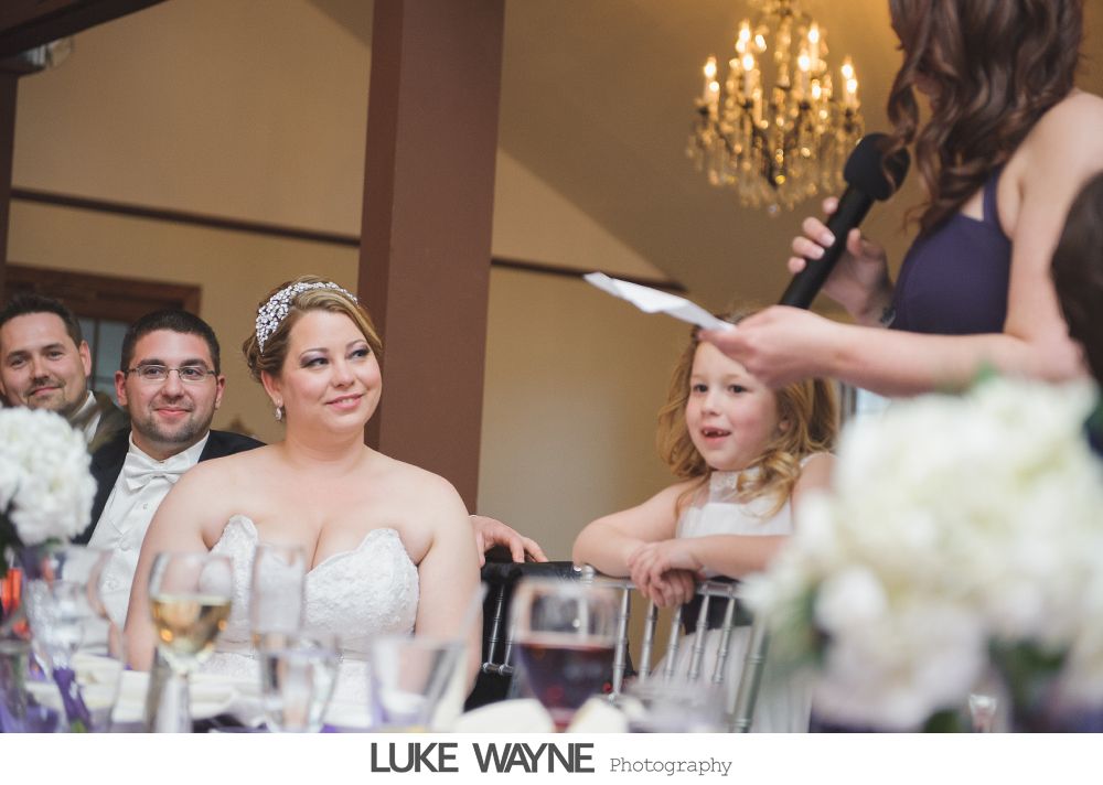 Bride smiling during a wedding toast, with flower girl and guests in a room with a chandelier.