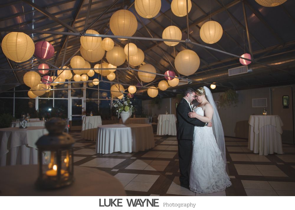 Newlyweds embrace in a well-lit reception hall with paper lanterns, surrounded by tables.