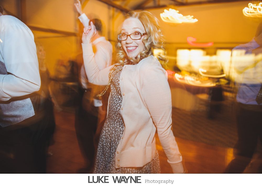 Woman with glasses dances at a party; arms raised, smiling. Yellowish lighting.