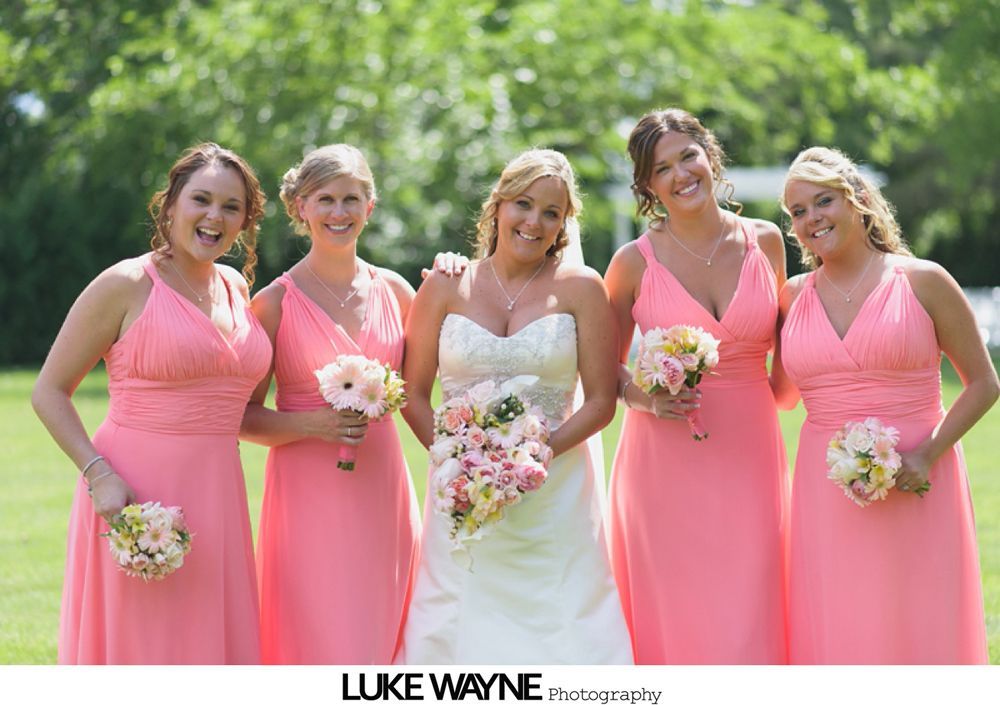 Bridesmaids in pastel dresses pose with bride outdoors near a building with lush green trees.