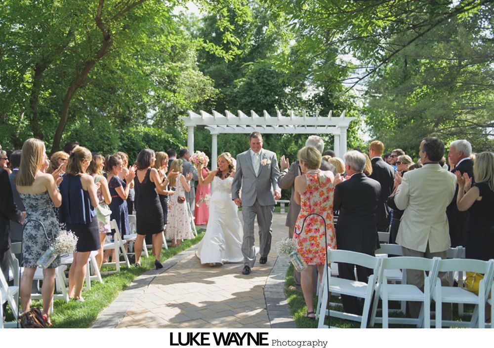 Bride walks down the aisle with her father at a wedding. Guests seated on either side of the aisle. Venue decorated with hanging lanterns.