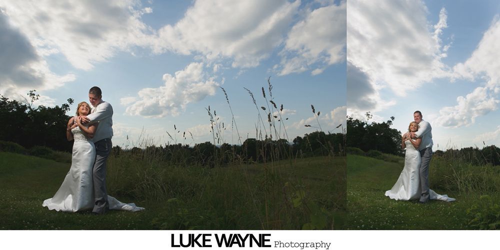 Wedding party posing in a field. People in formal wear with bouquets, smiling. Green field, cloudy sky.