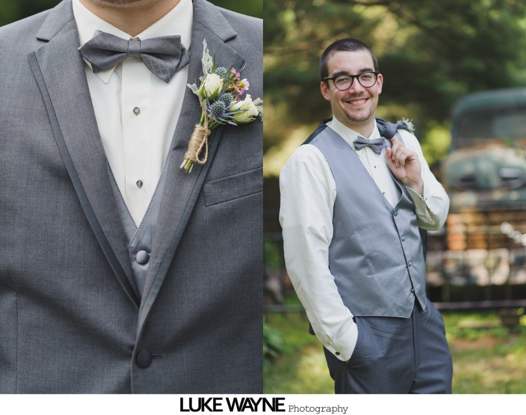 Man in gray suit with bow tie and boutonniere, smiling, outside near an old vehicle.