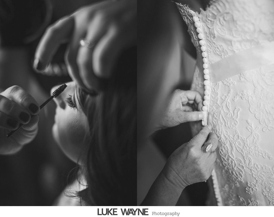 Black and white photo of a person getting makeup applied and the back of a wedding dress being buttoned up.