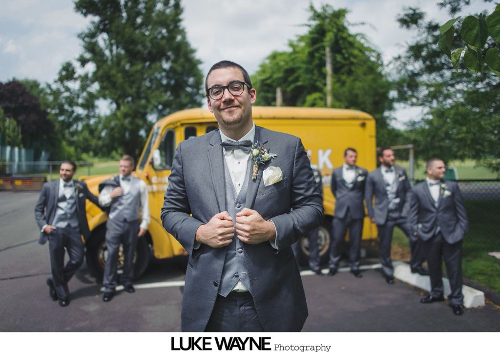 Groom in gray suit and bow tie smiles, posed with groomsmen, in front of yellow truck.