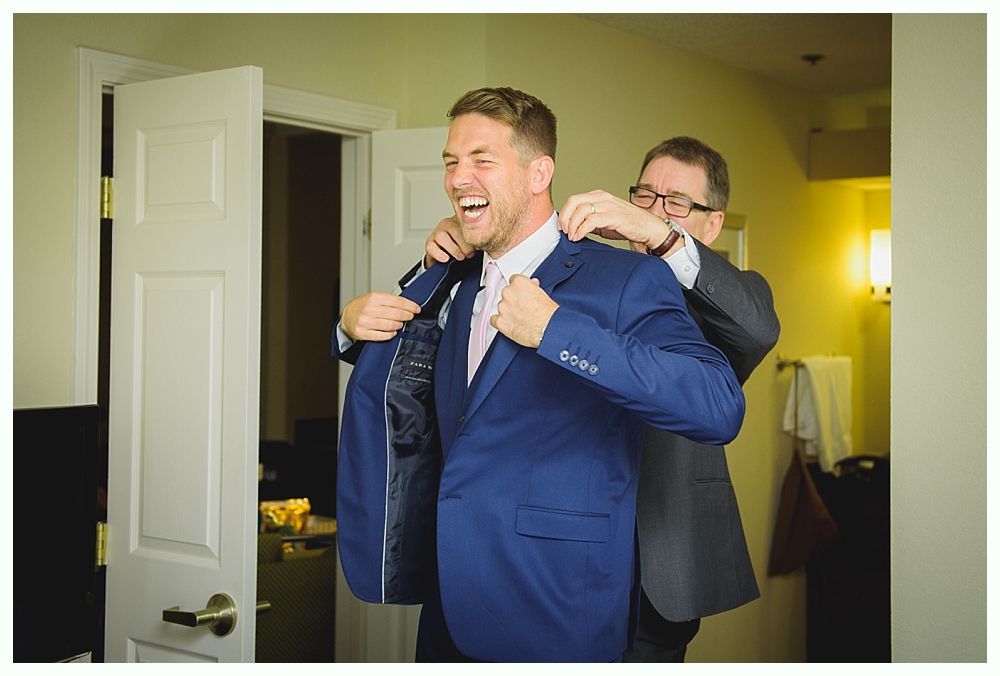 Man laughs as another adjusts his blue suit jacket in a room with a closed door.