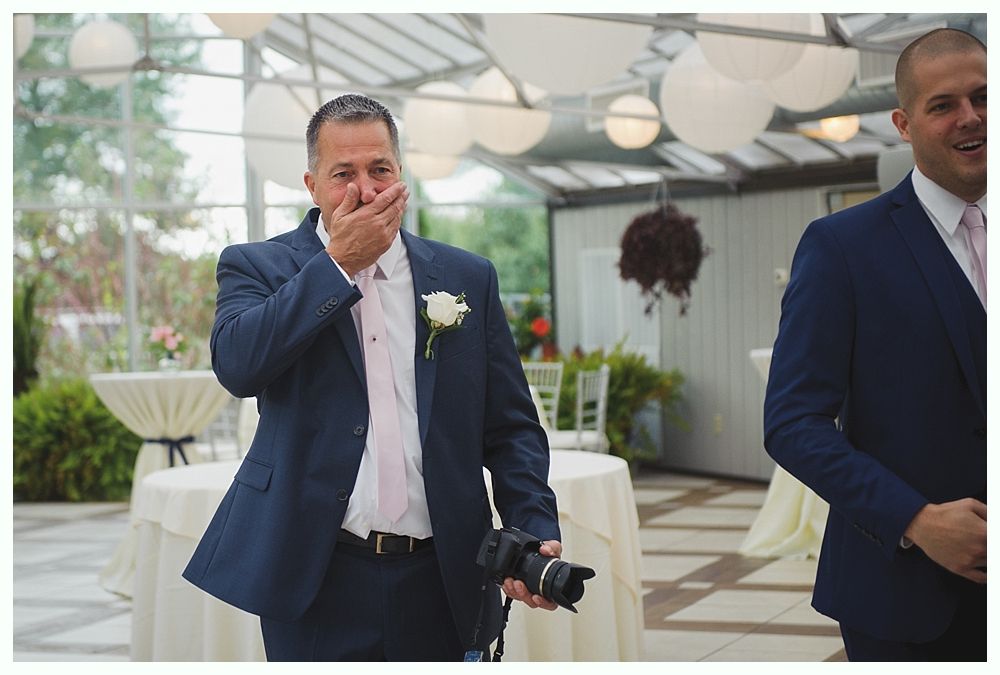 Man in blue suit, emotional, covers mouth, holds camera, wedding setting.