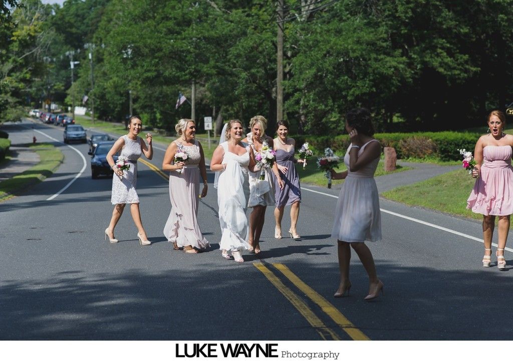 Bridesmaids in pastel dresses and bride in white, walking down a road, holding bouquets, sunny day.