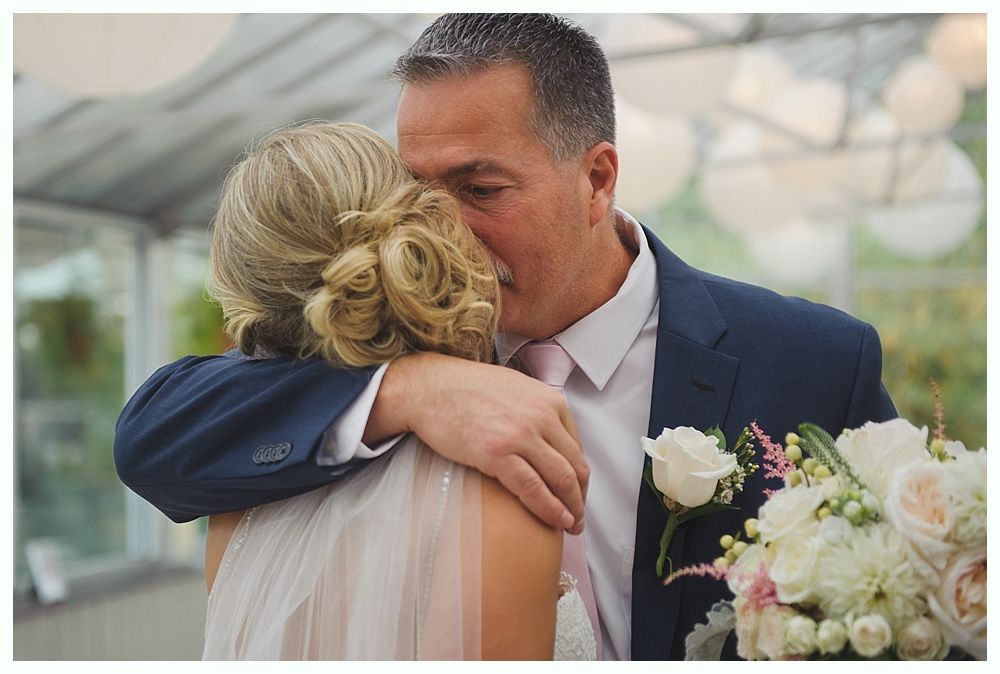 Man in suit hugs bride, touching her cheek, floral bouquet in hand.