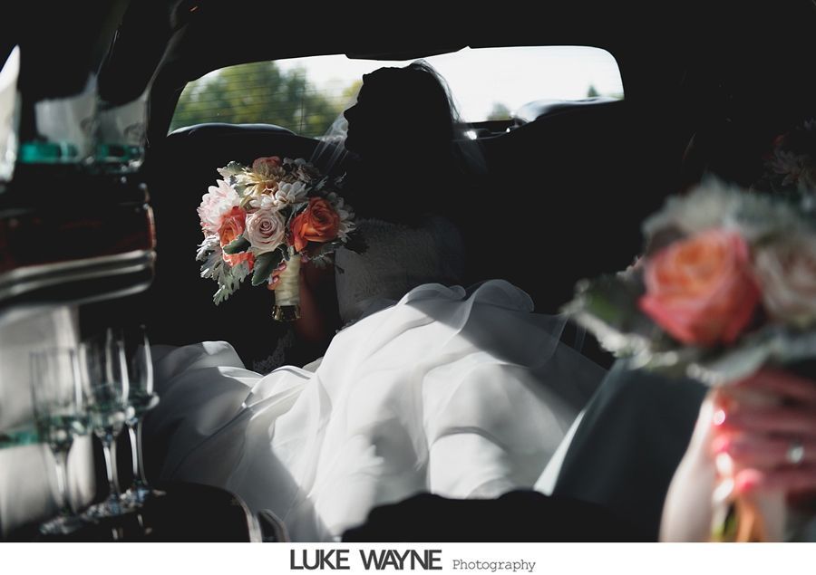 Bride in white dress holding bouquet inside limousine; sunlight streams in.