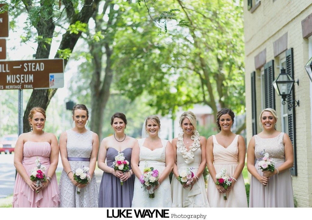 Bridesmaids in pastel dresses hold bouquets, posing outside a building next to a sign for the Hill-Stead Museum.