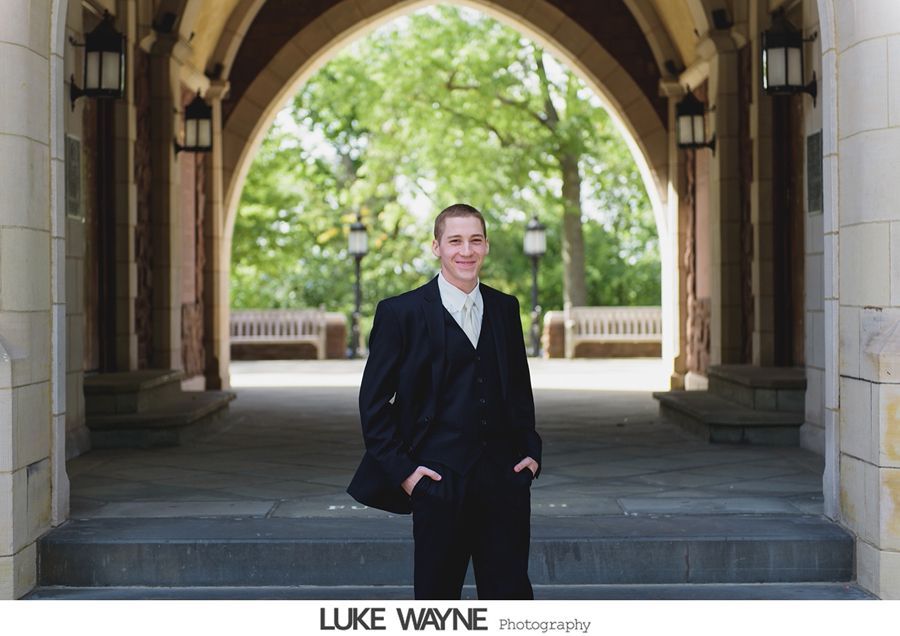 Man in suit stands under an arched walkway with hands in pockets; trees in background.