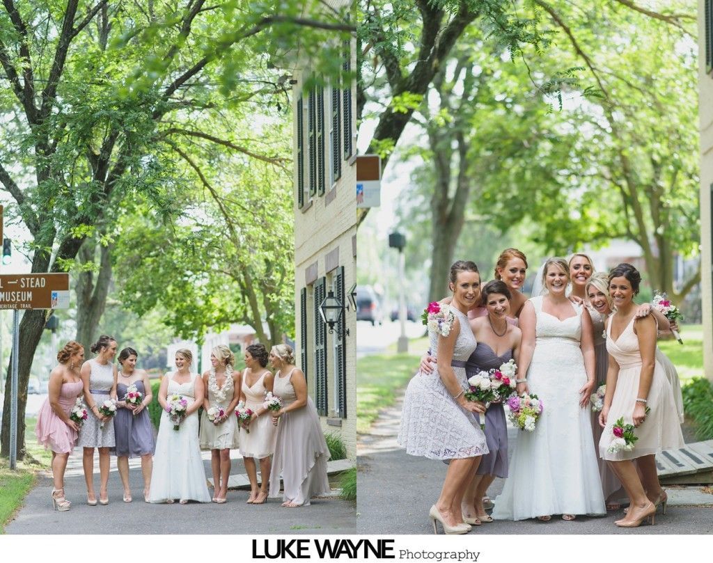 Bridesmaids in pastel dresses pose with bride outdoors near a building with lush green trees.