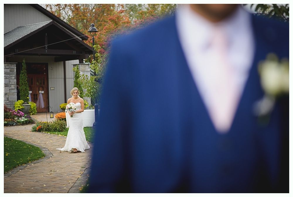 Bride walking toward groom, outdoor wedding. Groom in blue suit, pink tie. Building in background, fall foliage.