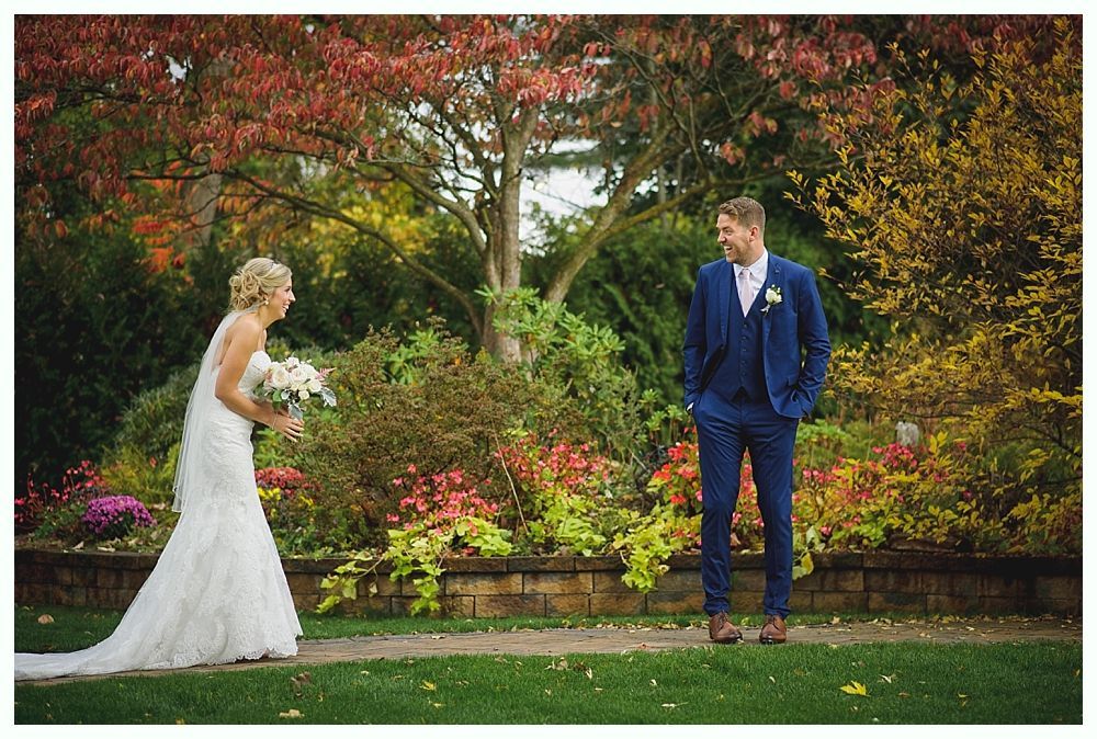 Bride laughs, approaches groom in a blue suit, holding bouquet. Fall foliage in background.