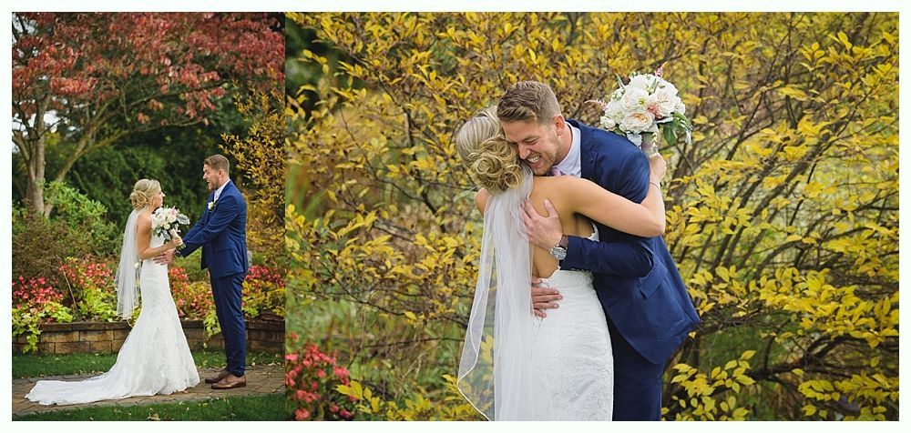 Newlyweds embrace in a garden. The bride wears white, groom in a blue suit. Lush trees with colorful foliage in the background.