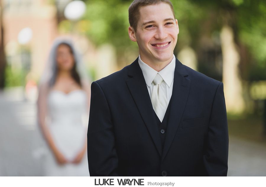 Groom smiling, wearing a suit, waiting for the bride in a street setting. Bride blurred in background.