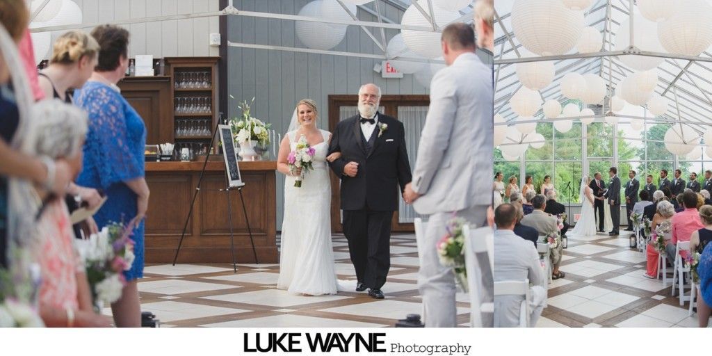 Bride walks down the aisle with her father at a wedding. Guests seated on either side of the aisle. Venue decorated with hanging lanterns.