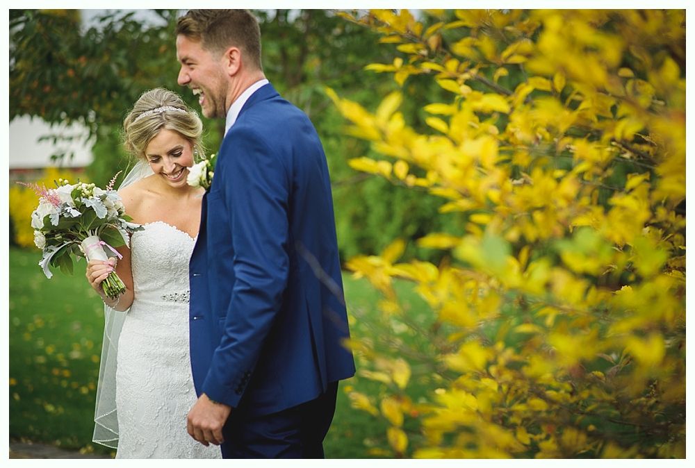 Bride and groom laughing outdoors; bride in white dress, holding bouquet, groom in blue suit.