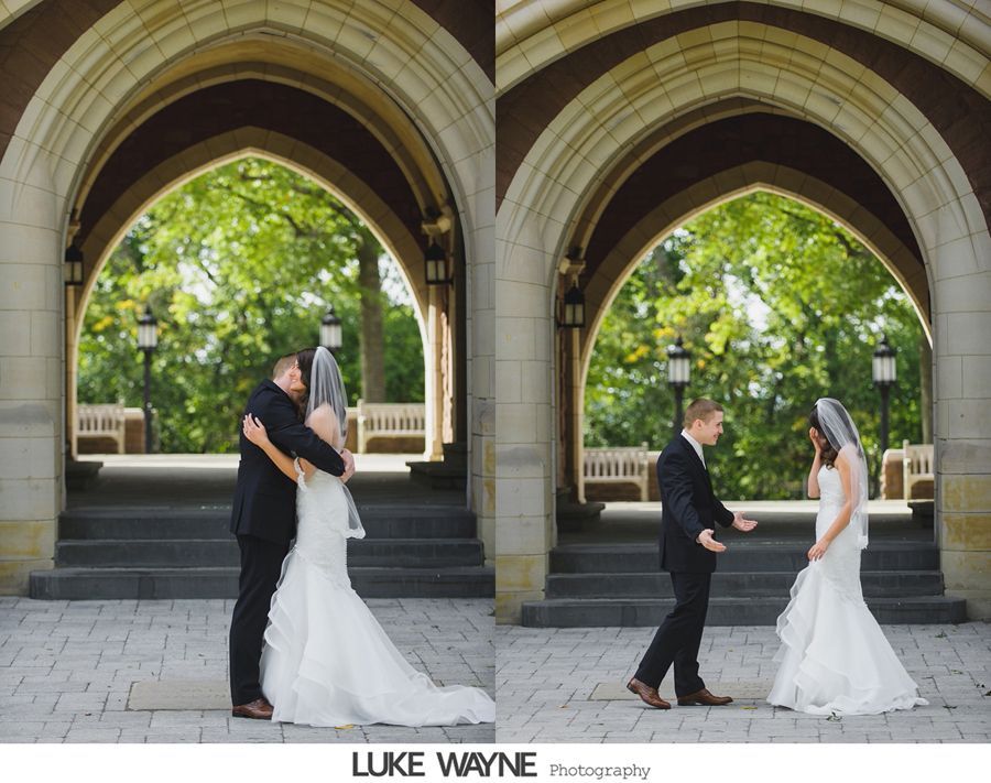 Bride and groom embrace, then look at each other. They stand under an archway, steps behind them, and trees beyond.