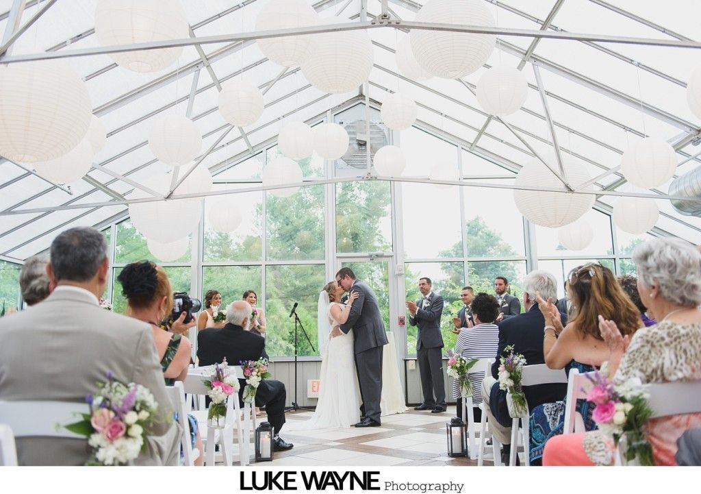 Couple kissing at their wedding ceremony inside a glass-roofed structure, surrounded by guests.