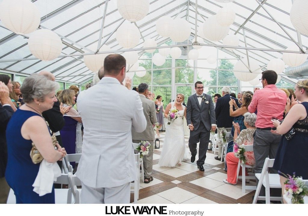 Newlyweds walking down the aisle after a wedding ceremony, guests clapping. Glass enclosed building, white paper lanterns overhead.