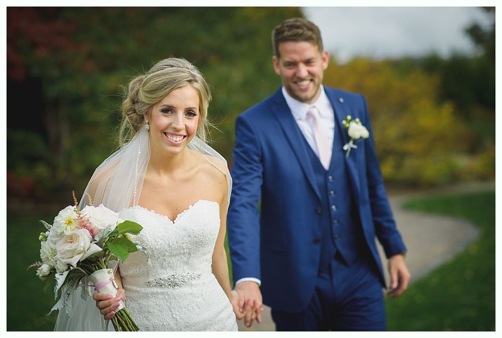 Bride and groom smiling, walking outside. Bride holds flowers, groom wears a blue suit.
