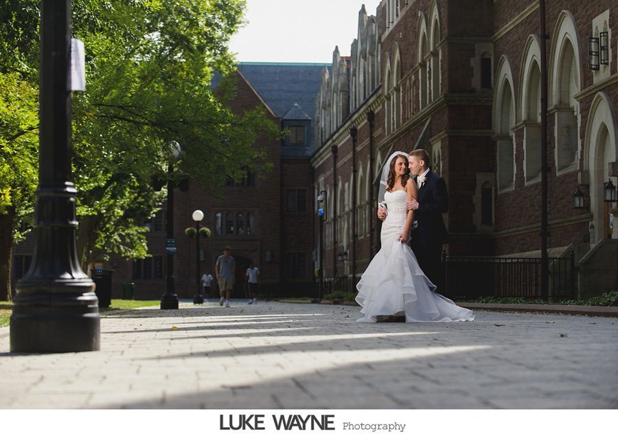 Bride and groom pose on brick path. They are near a brick building with trees.