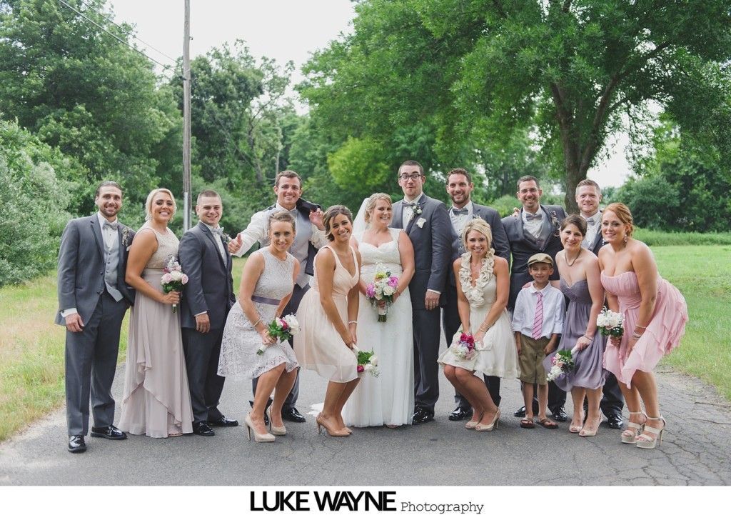 Wedding party posing on a road, with the bride and groom at the center. Some are smiling, others are making silly faces.