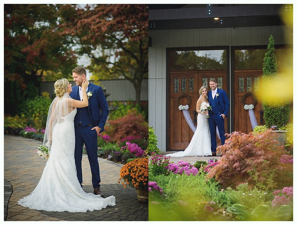Bride and groom pose outdoors. She wears a white gown, and he wears a blue suit. Flowers and greenery surround them.