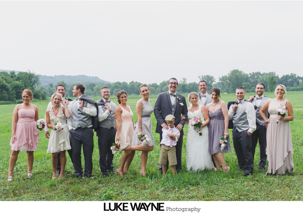 Wedding party posing in a field. People in formal wear with bouquets, smiling. Green field, cloudy sky.