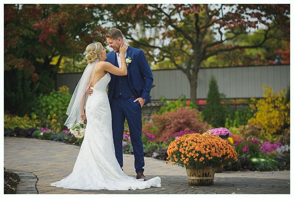 Bride in white dress touches groom's face, both smiling. They stand in a garden.