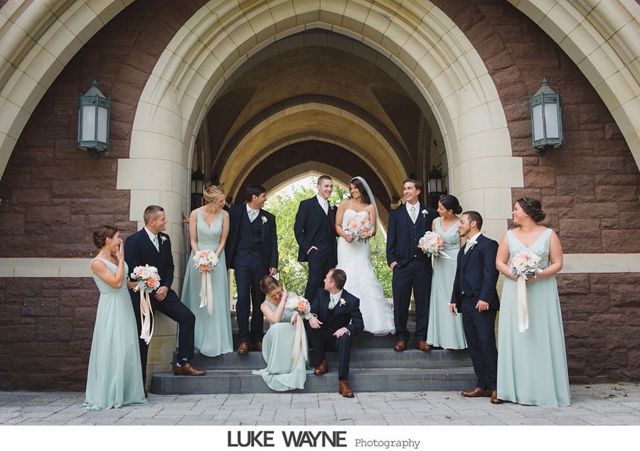 Wedding party posing under a brick archway. Bride and groom in center, flanked by bridesmaids in light green dresses and groomsmen in navy suits.