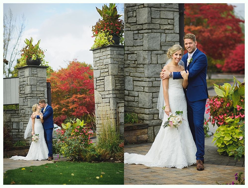 Bride and groom pose near stone pillars and autumn foliage. The bride wears white lace, the groom a blue suit.