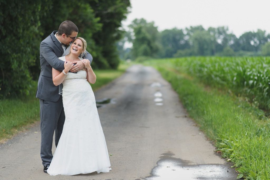 Bride and groom embrace on a country road, laughing, with green field and trees in the background.