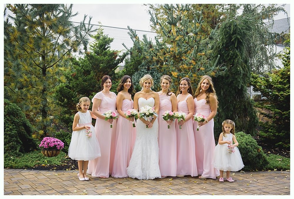 Bride in white gown with bridesmaids in pink dresses, posed outdoors with flower girls.