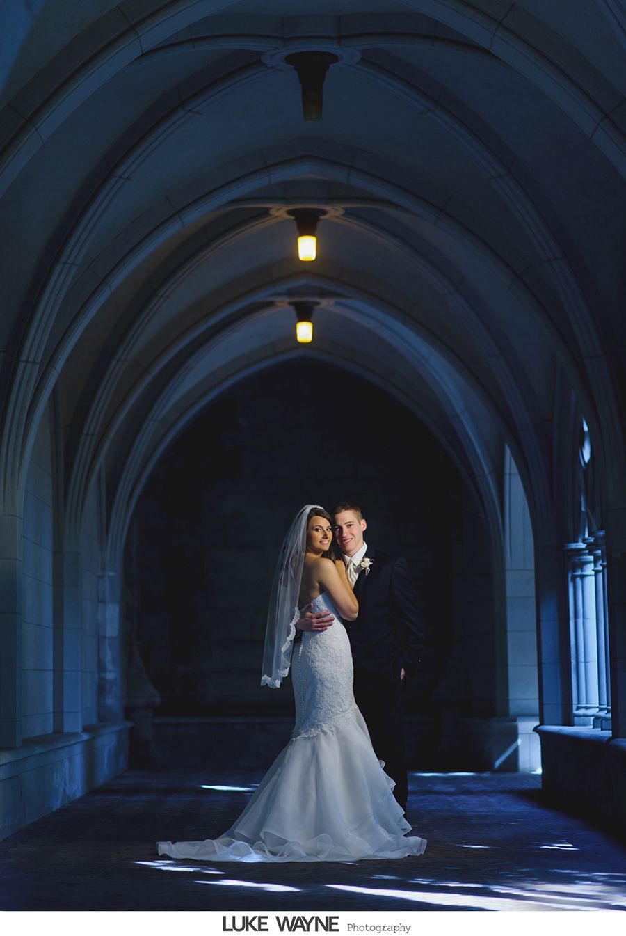 Bride and groom stand in a dark arched hallway, wedding attire. Soft lighting above them.