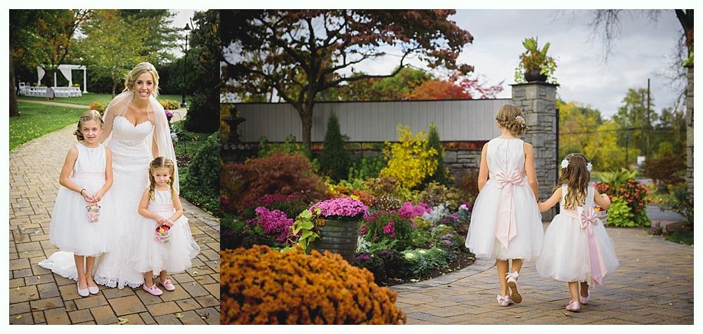 Bride with flower girls, walking on a path. They are wearing white dresses. Colorful flower garden and trees in the background.