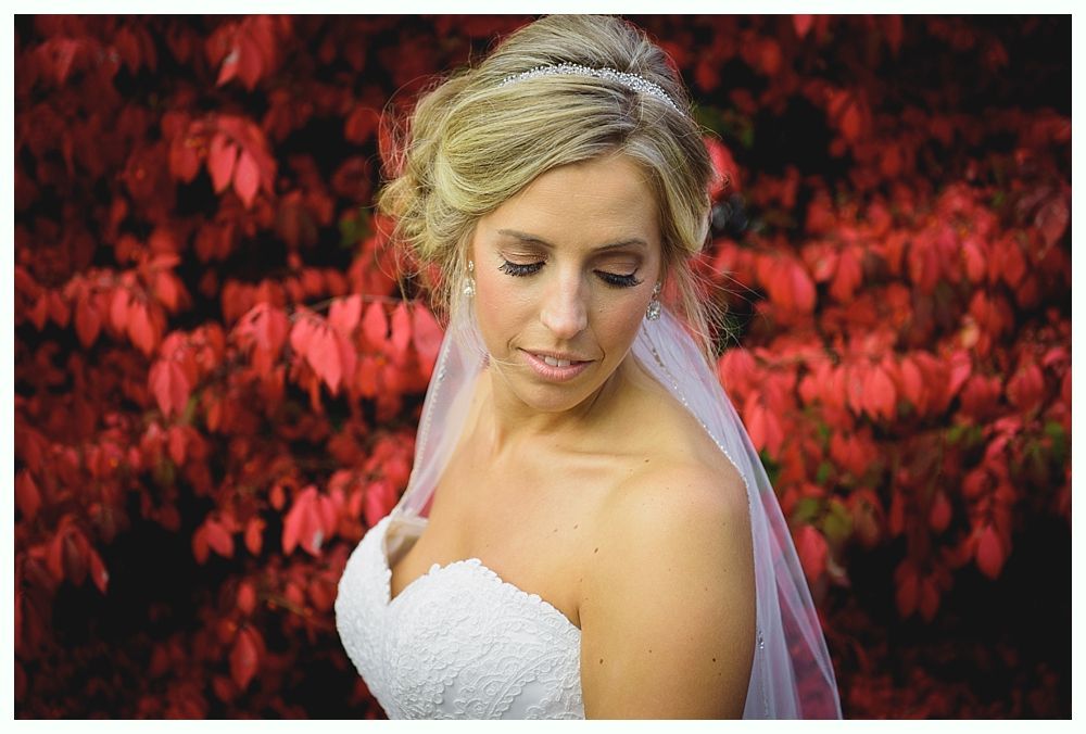 Bride in white dress with veil, looking down. Red foliage background.