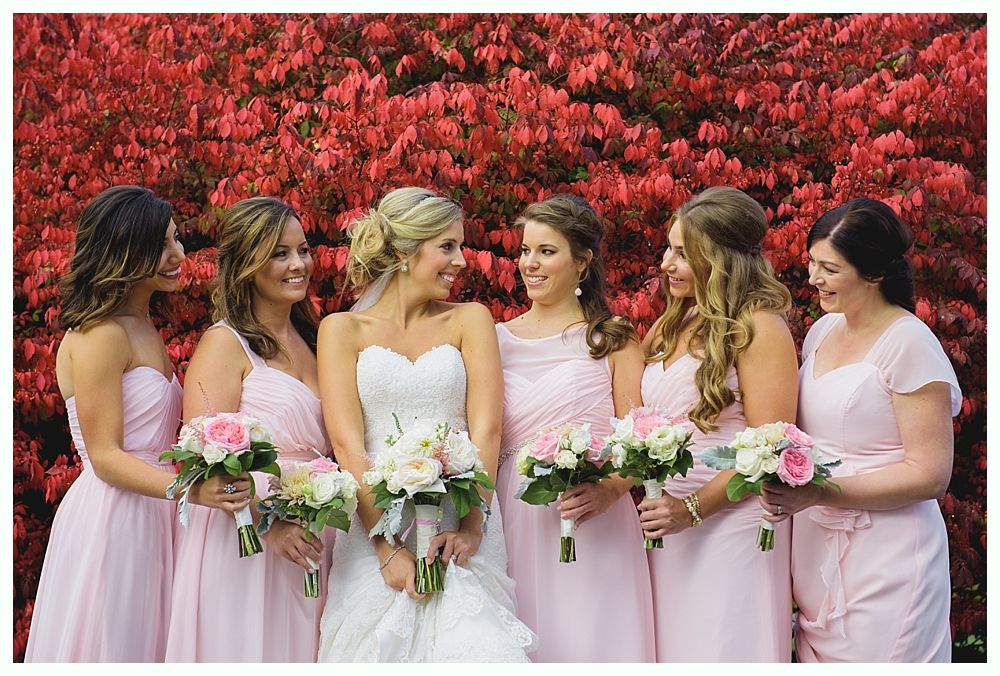 Bride in white gown with bridesmaids in pink dresses, holding bouquets, red foliage background.