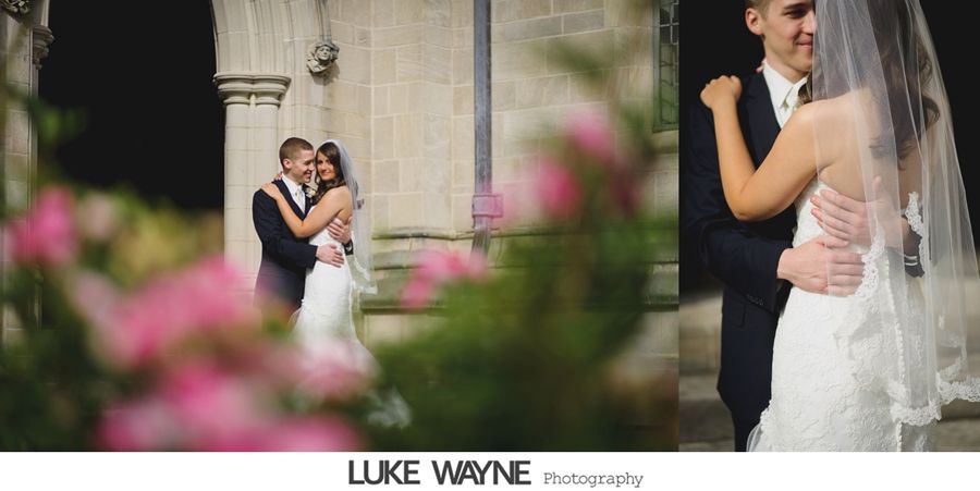 Newlyweds embrace, posing for a photo. Bride in a lace gown, groom in a dark suit, flowers in foreground, stone building in background.