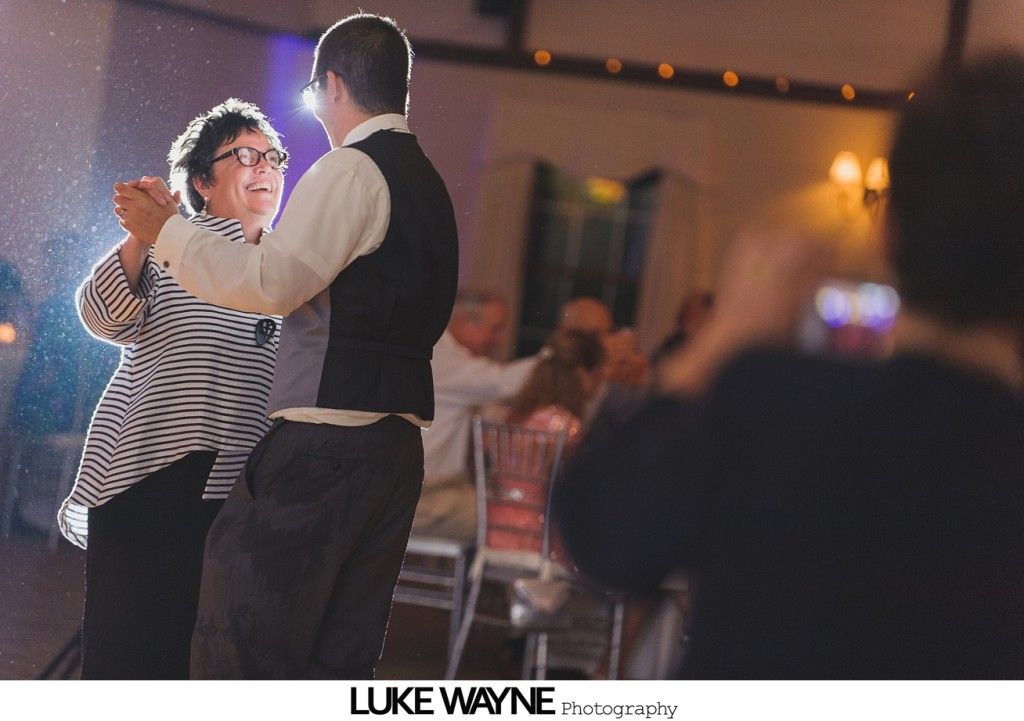 Man dances with a woman at a reception; indoor setting. Woman wears glasses and striped top. Person takes photo.