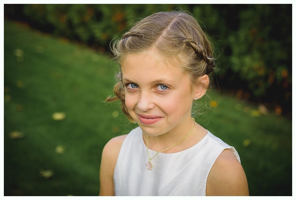 Young girl with braided hair and blue eyes, smiling. Wearing a white dress, set outdoors with green grass.