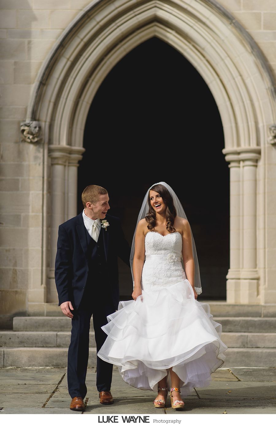 Bride and groom smiling, leaving church entrance. Bride in white dress and veil, groom in dark suit.