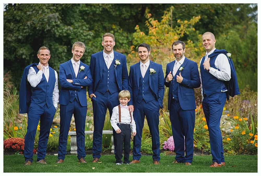 Groomsmen and a young boy in navy suits pose outdoors. Flowers and greenery are in the background.