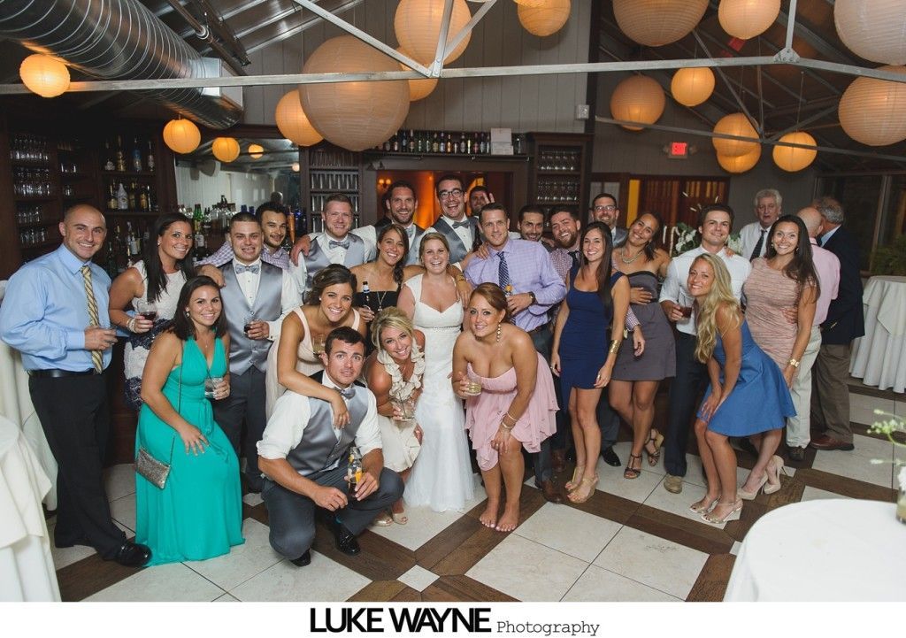 Group photo at a wedding reception. Bride and groom centered, surrounded by guests in formal attire.