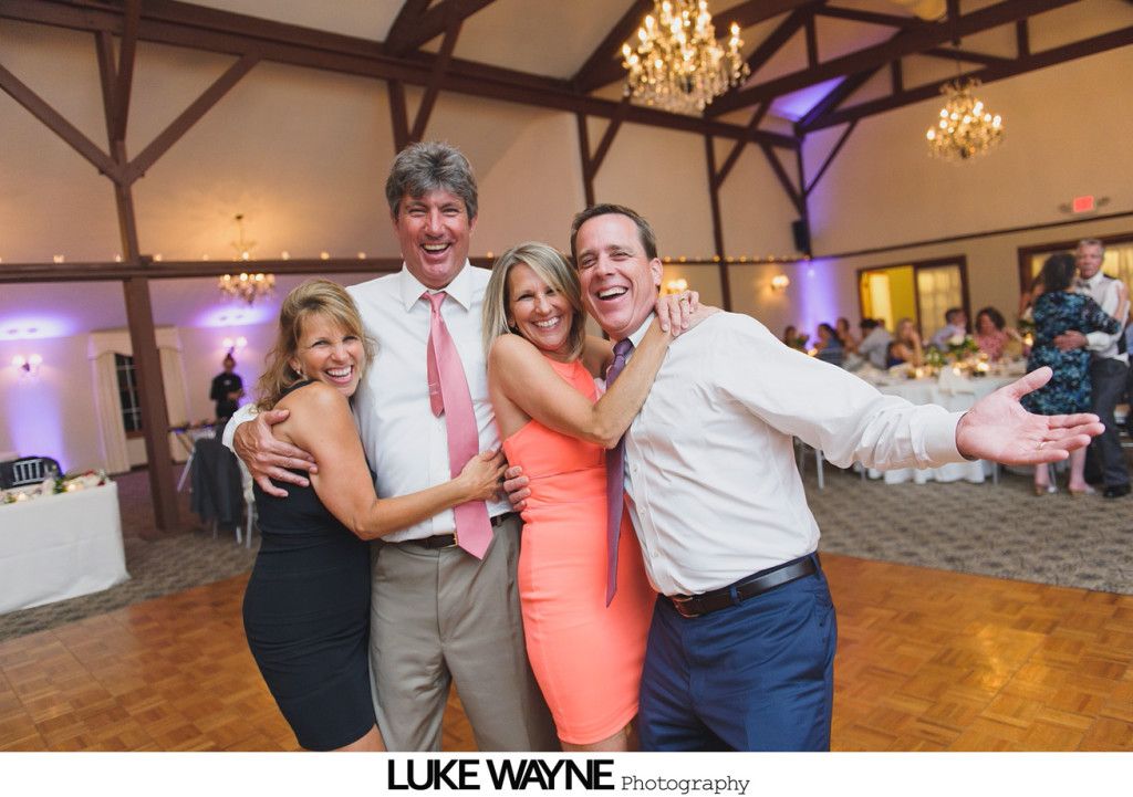 Four people smiling and embracing on a wooden dance floor in a reception hall.