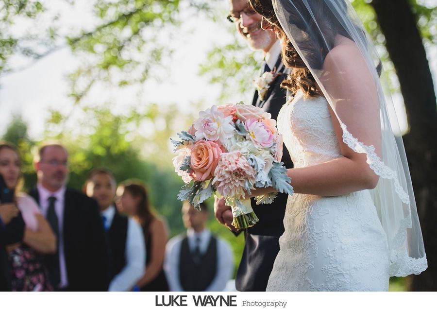 Bride holding bouquet walking towards ceremony, with veil, other guests and trees in the background.
